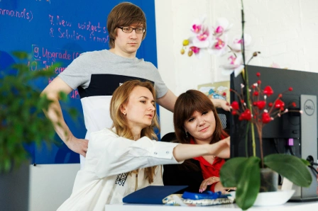 Three people working at a computer, with a whiteboard in the background and plants in the foreground.