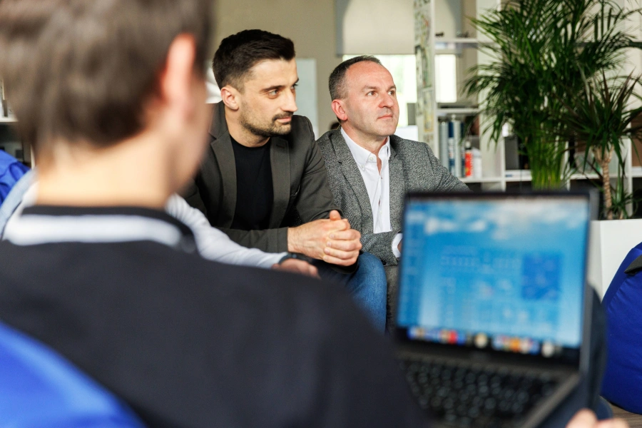 Two men sit attentively in an informal setting, while a person in the foreground uses a laptop. A large plant and bookshelves are in the background, creating a relaxed office atmosphere.