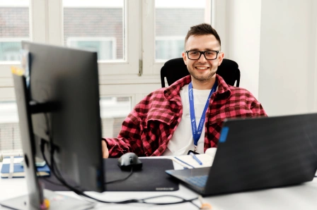 A person wearing glasses and a red checkered shirt sits at a desk with a computer monitor and a laptop. He is smiling and there is a window in the background.